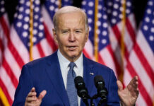 Man speaking in front of American flags and microphones