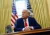 A man in a suit sitting at a desk in the Oval Office with American flags in the background