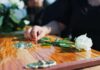 A hand placing a white rose on a wooden casket during a funeral service